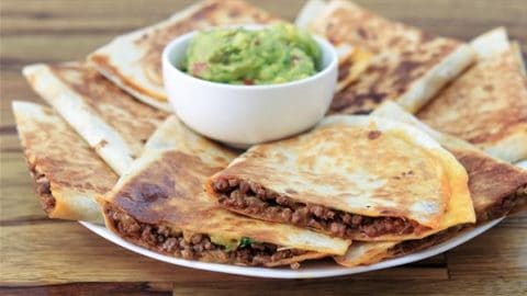 A plate of golden-brown quesadillas, filled with seasoned ground beef, sits on a wooden surface. The quesadillas are neatly arranged around a small white bowl of guacamole placed in the center of the plate.