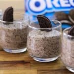 Four glasses filled with cookies and cream mousse are neatly arranged in a row on a wooden surface. Each glass is topped with a half Oreo cookie. In the background, there is a blurred Oreo cookie package.