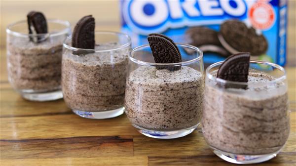 Four glasses filled with cookies and cream mousse are neatly arranged in a row on a wooden surface. Each glass is topped with a half Oreo cookie. In the background, there is a blurred Oreo cookie package.