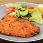 A grey plate with a breaded and fried schnitzel on one side and a colorful salad with lettuce, cucumber, tomato, bell peppers, and a lemon wedge on the other side. The plate is on a wooden surface.