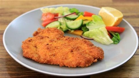 A grey plate with a breaded and fried schnitzel on one side and a colorful salad with lettuce, cucumber, tomato, bell peppers, and a lemon wedge on the other side. The plate is on a wooden surface.