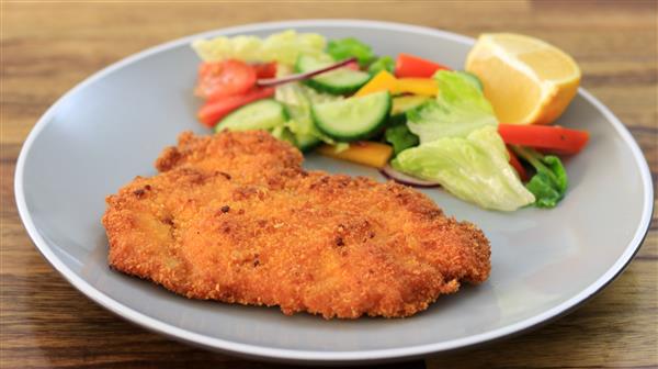 A grey plate with a breaded and fried schnitzel on one side and a colorful salad with lettuce, cucumber, tomato, bell peppers, and a lemon wedge on the other side. The plate is on a wooden surface.