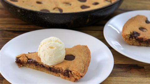 A slice of chocolate chip cookie pizza topped with a scoop of vanilla ice cream is served on a white plate. Another similar slice is on a separate plate in the background, with the rest of the cookie pizza visible in a skillet. The scene is set on a wooden table.