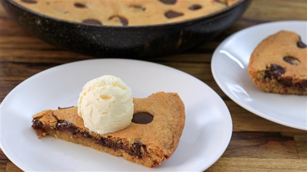 A slice of chocolate chip cookie pizza topped with a scoop of vanilla ice cream is served on a white plate. Another similar slice is on a separate plate in the background, with the rest of the cookie pizza visible in a skillet. The scene is set on a wooden table.