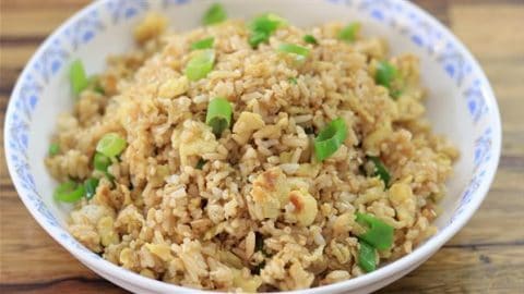 A bowl filled with golden brown fried rice, garnished with chopped green onions. The rice appears to have small pieces of scrambled egg mixed in, and the bowl is placed on a wooden table. The rim of the bowl has a blue floral pattern.