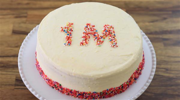 A round cake with smooth white frosting is displayed on a white platter. The top of the cake has the text "1M" written in multi-colored sprinkles, and the bottom edge of the cake is also decorated with a border of the same sprinkles.