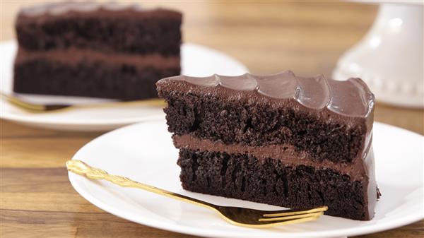 Two slices of rich chocolate cake with thick, glossy chocolate frosting are placed on white plates. Each plate includes a golden fork, and the chocolate cake layers appear moist and decadent. The background is a light wooden surface.