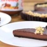 A slice of chocolate tart with a crumbly dark crust and sprinkled with chopped nuts on top sits on a white plate. In the background, a whole tart and a jar of Nutella are visible on a wooden surface.