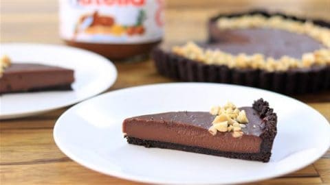 A slice of chocolate tart with a crumbly dark crust and sprinkled with chopped nuts on top sits on a white plate. In the background, a whole tart and a jar of Nutella are visible on a wooden surface.