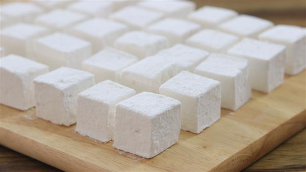 A wooden board holds rows of white, fluffy, cube-shaped marshmallows neatly arranged in a grid pattern.