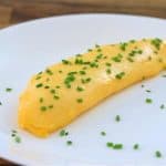 A perfectly cooked, golden yellow omelette garnished with finely chopped chives is plated on a plain white dish. The wooden tabletop serves as a rustic background, adding warmth to the presentation.