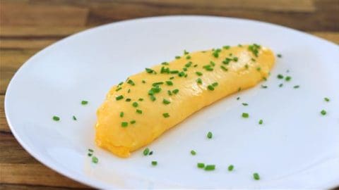 A perfectly cooked, golden yellow omelette garnished with finely chopped chives is plated on a plain white dish. The wooden tabletop serves as a rustic background, adding warmth to the presentation.