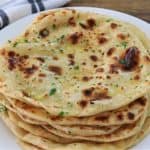 A plate of homemade naan bread topped with fresh herbs sits on a wooden table. The naan is golden brown with charred spots, and a folded kitchen towel is visible in the background.