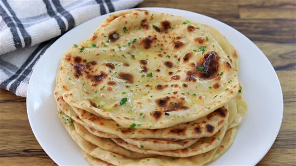 A plate of homemade naan bread topped with fresh herbs sits on a wooden table. The naan is golden brown with charred spots, and a folded kitchen towel is visible in the background.