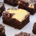 Close-up of a batch of marbled chocolate and cream cheese brownies, cut into individual square pieces and arranged on a white surface. The brownies feature a rich, dark chocolate base with a creamy, light yellow cream cheese swirl on top.