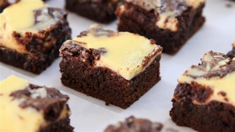 Close-up of a batch of marbled chocolate and cream cheese brownies, cut into individual square pieces and arranged on a white surface. The brownies feature a rich, dark chocolate base with a creamy, light yellow cream cheese swirl on top.