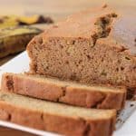 A loaf of banana bread partly sliced on a white rectangular plate. Two ripe bananas are in the background, resting on a wooden surface. The bread appears moist and dense, with a cracked top crust.