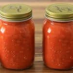 Two jars filled with homemade tomato sauce sit on a wooden surface, accompanied by three fresh tomatoes placed around them. The jars have golden lids with decorative designs on top, and the tomato sauce inside appears rich and chunky.