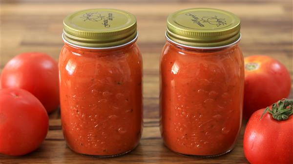 Two jars filled with homemade tomato sauce sit on a wooden surface, accompanied by three fresh tomatoes placed around them. The jars have golden lids with decorative designs on top, and the tomato sauce inside appears rich and chunky.