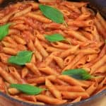 A pot of cooked penne pasta mixed with tomato sauce and garnished with fresh basil leaves on a wooden surface. The pasta is evenly coated in the sauce, and the vibrant green basil leaves are placed on top for decoration.