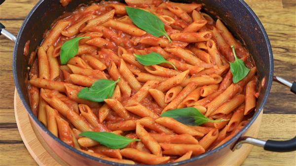 A pot of cooked penne pasta mixed with tomato sauce and garnished with fresh basil leaves on a wooden surface. The pasta is evenly coated in the sauce, and the vibrant green basil leaves are placed on top for decoration.