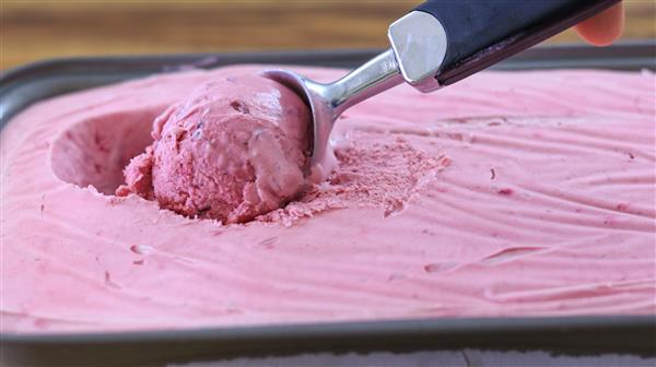 A close-up image of a hand using an ice cream scoop to serve a portion of pink, creamy ice cream from a rectangular container. The ice cream has a smooth texture with light ridges from the scoop. The background is slightly blurred.