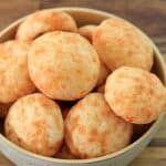 A bowl filled with several Brazilian cheese breads, known as "pão de queijo," is placed on a wooden surface. The golden, round, and slightly crispy breads have a smooth texture dotted with small bubble-like spots, indicating a light, airy interior.