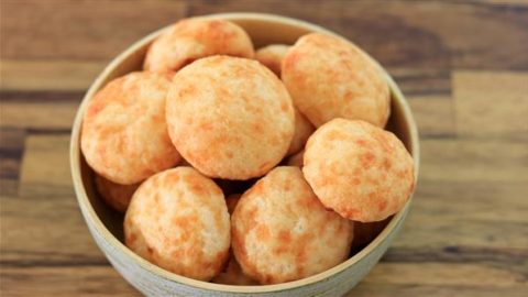 A bowl filled with several Brazilian cheese breads, known as "pão de queijo," is placed on a wooden surface. The golden, round, and slightly crispy breads have a smooth texture dotted with small bubble-like spots, indicating a light, airy interior.