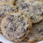 A white plate filled with cookies sits on a wooden surface. The cookies are light-colored with visible chunks of cookies 'n' cream embedded in them, giving a slightly speckled appearance. They appear soft and chewy.