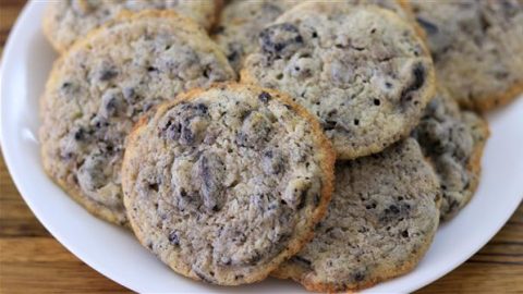 A white plate filled with cookies sits on a wooden surface. The cookies are light-colored with visible chunks of cookies 'n' cream embedded in them, giving a slightly speckled appearance. They appear soft and chewy.