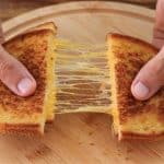 A pair of hands pulling apart a grilled cheese sandwich on a wooden plate, revealing the gooey, melted cheese stretching between the two halves. The sandwich is golden brown and crispy.