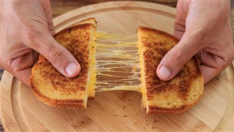 A pair of hands pulling apart a grilled cheese sandwich on a wooden plate, revealing the gooey, melted cheese stretching between the two halves. The sandwich is golden brown and crispy.