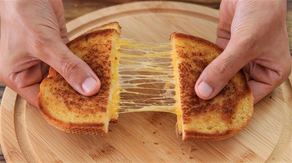 A pair of hands pulling apart a grilled cheese sandwich on a wooden plate, revealing the gooey, melted cheese stretching between the two halves. The sandwich is golden brown and crispy.