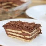 A piece of layered chocolate biscuit cake is served on a white plate. The cake has alternating layers of biscuits and chocolate cream, topped with chocolate shavings. In the background, there is a glass dish containing more of the same cake.