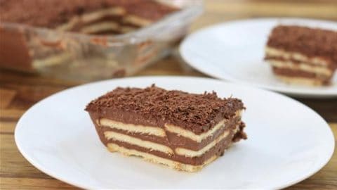 A piece of layered chocolate biscuit cake is served on a white plate. The cake has alternating layers of biscuits and chocolate cream, topped with chocolate shavings. In the background, there is a glass dish containing more of the same cake.