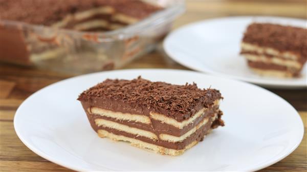 A piece of layered chocolate biscuit cake is served on a white plate. The cake has alternating layers of biscuits and chocolate cream, topped with chocolate shavings. In the background, there is a glass dish containing more of the same cake.