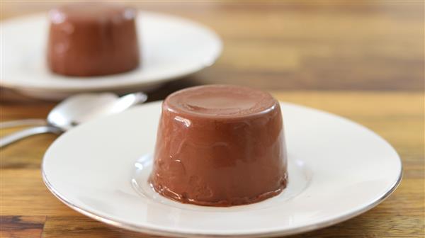 Two servings of chocolate panna cotta are presented on white plates. The panna cottas have a smooth, rich texture and are accompanied by spoons. The scene is set on a wooden table.