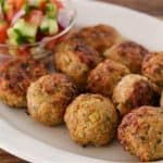 An oval white plate holds ten golden-brown meatballs. A small, clear bowl filled with a colorful vegetable salad—consisting of diced tomatoes, cucumbers, and onions—is placed at the edge of the platter. The background is a wooden surface.