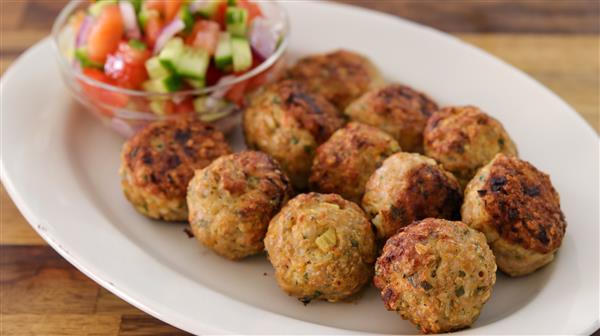 An oval white plate holds ten golden-brown meatballs. A small, clear bowl filled with a colorful vegetable salad—consisting of diced tomatoes, cucumbers, and onions—is placed at the edge of the platter. The background is a wooden surface.