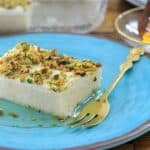 A slice of white dessert topped with chopped pistachios sits on a blue plate. There is a small golden dessert fork next to it on the plate. In the background, a glass of tea on a saucer is visible.