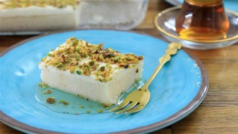 A slice of white dessert topped with chopped pistachios sits on a blue plate. There is a small golden dessert fork next to it on the plate. In the background, a glass of tea on a saucer is visible.