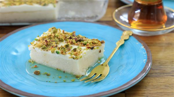 A slice of white dessert topped with chopped pistachios sits on a blue plate. There is a small golden dessert fork next to it on the plate. In the background, a glass of tea on a saucer is visible.