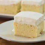 A slice of tres leches cake sits on a white plate. The cake is topped with a layer of fluffy white frosting, and the moist, pale yellow base is visible. In the background, more slices of cake are displayed on a rectangular serving dish.