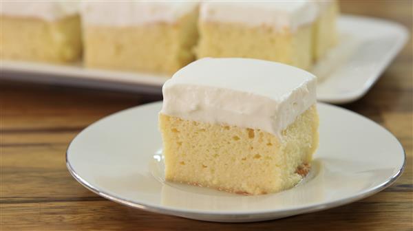 A slice of tres leches cake sits on a white plate. The cake is topped with a layer of fluffy white frosting, and the moist, pale yellow base is visible. In the background, more slices of cake are displayed on a rectangular serving dish.