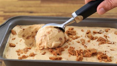 A hand is using an ice cream scoop to serve caramel ice cream from a metal tray. The ice cream is topped with crushed cookie pieces and appears creamy and rich. The background is a wooden surface.