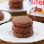 A plate on a wooden table holds three stacked chocolate cookies. In the background, another plate with more cookies and a jar of Nutella are visible.