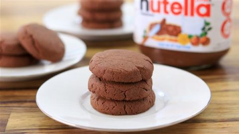 A plate on a wooden table holds three stacked chocolate cookies. In the background, another plate with more cookies and a jar of Nutella are visible.