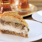 A slice of layered dessert featuring a golden, flaky crust filled with creamy and nutty layers, served on a white plate. In the background, there are traditional glasses on a tray, likely used for serving tea.