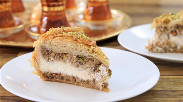 A slice of layered dessert featuring a golden, flaky crust filled with creamy and nutty layers, served on a white plate. In the background, there are traditional glasses on a tray, likely used for serving tea.