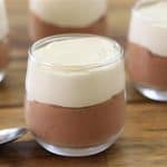 Small glass jars filled with a dessert, consisting of a bottom layer of chocolate mousse and a top layer of vanilla mousse, are arranged on a wooden surface. Two spoons are placed among the jars.